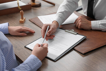 Notary pointing at document and client putting signature at wooden table indoors, closeup