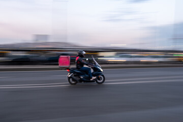 Obraz premium Urban Fast Food Delivery Person on Scooter Racing Through Streets at Dusk: Motion Blur Captures Speed, Efficiency, and Dynamic City Transportation