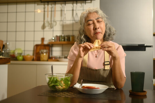 A Senior Asian Woman Savoring a Homemade Sandwich in Cozy Kitchen Setting