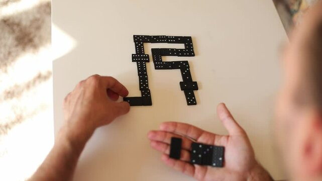 A close-up shot of a hand arranging black dominoes on a white surface, forming a creative pattern. The domino pieces are neatly laid out, and the blurred background adds focus to the action.