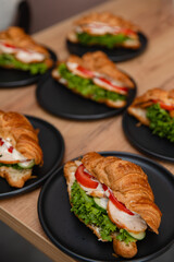 Tasty croissant sandwiches with fresh lettuce, tomato, cucumber, chicken, and sauce on black plates. Vertical food photography, ideal for breakfast, brunch, or catering presentations.