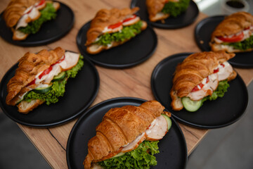 Delicious croissant sandwiches with lettuce, tomato, cucumber, chicken, and sauce on black plates, served on a wooden table. Perfect for breakfast, brunch, or catering presentations.