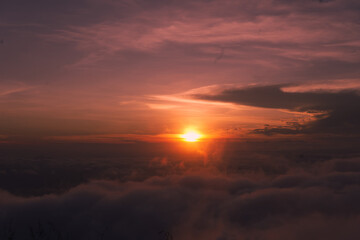 Golden sunrise lights up the sky with soft purple and orange hues above a blanket of clouds, creating a peaceful and dreamy view from the mountaintop.