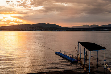A dock is on the water with a person standing on it. The water is calm and the sky is orange