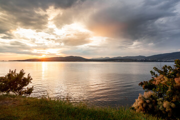 A beautiful sunset over a calm lake with a few trees in the background. The sky is filled with clouds, creating a moody atmosphere