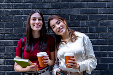 Two businesswomen are holding takeaway coffee, smiling, and enjoying their break near a brick wall