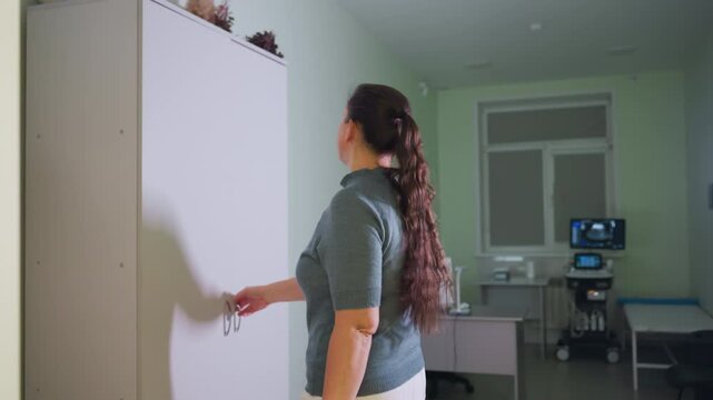 Beautiful tall female medical staff enters clinic room, walks directly to dresser, opens cabinet to retrieve uniform, then closes drawer behind her as she prepares to start professional work