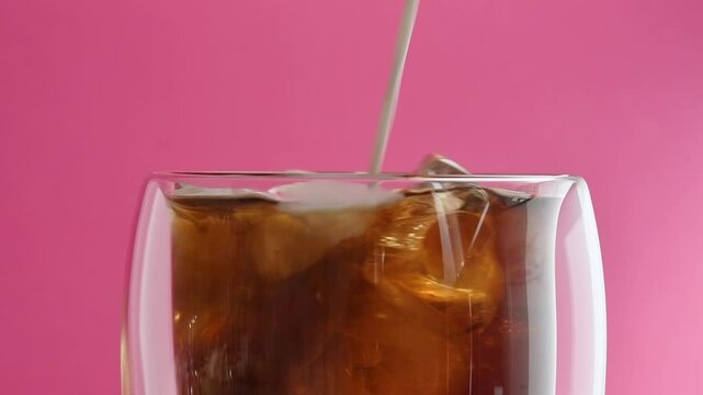 Cold brew with ice and creamy swirl slowly mixes in rotating glass against pink background, creating dreamy visual delight