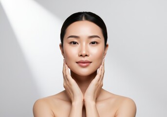 A modern beauty portrait of an Asian woman with her hair tied back and flawless radiant skin and gently holds her jawline with both hands