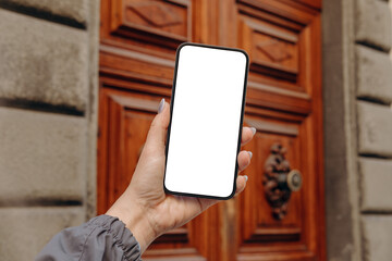 Mockup female  hand holding using smart phone with white blank screen POV at on the background of a house on the street in Europe real estate rent, mobile app design and advertising, online marketing	