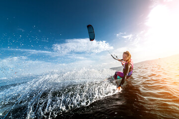 Kitesurfing girl in sexy wetsuit with kite in sky on board in blue sea riding on waves with water splash. Recreational activity, water sports, action, hobby and fun in summer. Kiteboarding sport