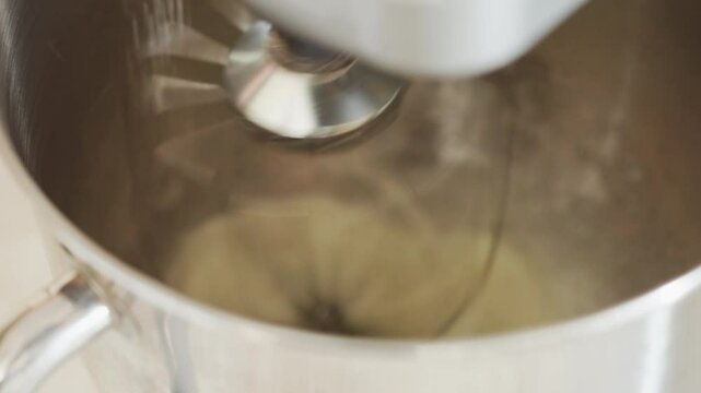 Close up of electric mixer in motion as flour is poured into stainless steel bowl during baking preparation showing dynamic spinning blade and smooth metallic texture in bright kitchen environment