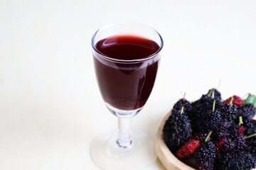 A glass of dark red mulberry juice sits next to a light wooden plate with ripe fruits on a cream table.