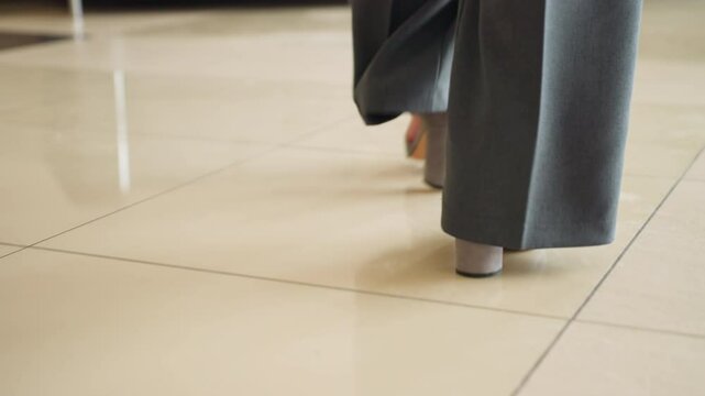 Close-up view of business professional in gray formal trousers and light socks walking across beige tiled floor in corporate office environment. Professional attire