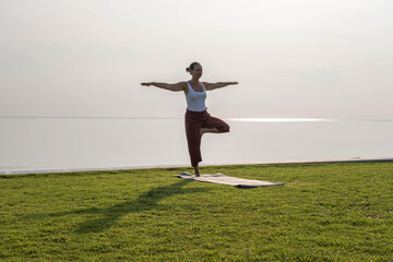 A woman gracefully performs yoga on a lush green shore, balancing in a serene pose as the gentle...
