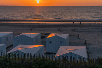 Fototapeta premium Beachside cabins glow under a vibrant sunset, reflecting warm hues. People enjoy a peaceful walk along the shoreline. Calm sea sets a serene backdrop on a picturesque evening. 