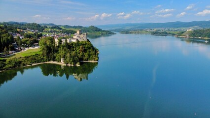 Aerial view of Niedzica Castle in Poland. The castle perched on a hill surrounded by lush greenery and a serene lake. The medieval structure features stone walls and towers