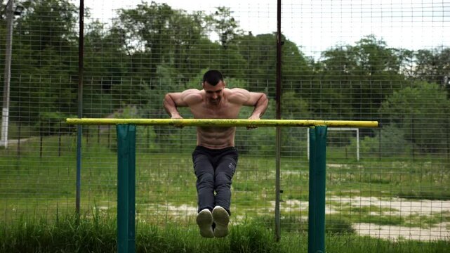 Hardy shirtless sportsman doing exercises on parallel bars at nature. Strong and muscular man working out at sports ground. Young athletic guy exercising outdoor. Concept of active healthy lifestyle