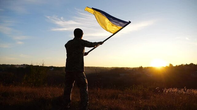 Ukrainian army man waving national banner against background of sunset. Young male soldier lifted flag at countryside. Victory against russian aggression. End of war between russia and Ukraine