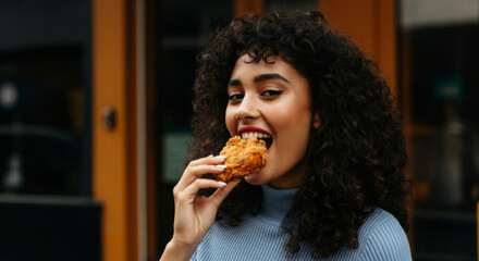 Young hispanic woman eating fried chicken outdoors in urban setting