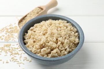 Tasty brown rice in bowl and raw grains on white wooden table, closeup
