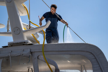 Luxury yacht being cleaned by a professional worker at the dock. Boat maintenance service in action at a marina with a bright summer sky and moored vessels.