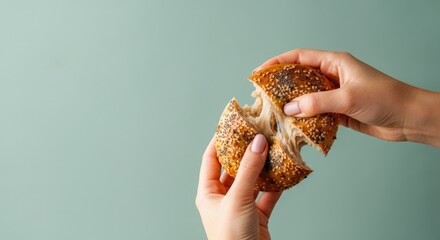 Hands tearing fresh sesame seed bagel on soft green background