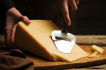 Woman cutting delicious cheese with slicer at wooden table against black background, closeup