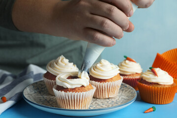 Woman squeezing whipped cream onto delicious carrot cupcakes at blue table, closeup