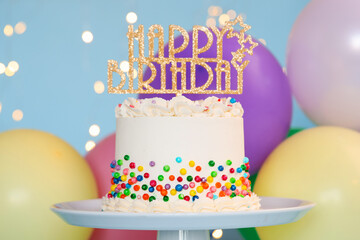 Delicious bento cake with colorful sprinkles and topper with words Happy Birthday on light blue background against blurred lights, closeup