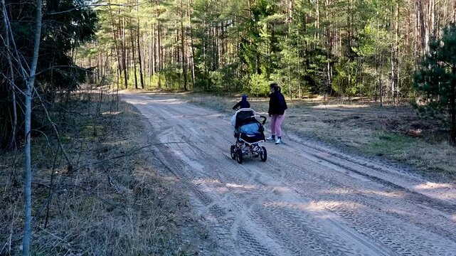 Mother assists child riding a bike on a forest trail with strollers nearby during a sunny day in spring