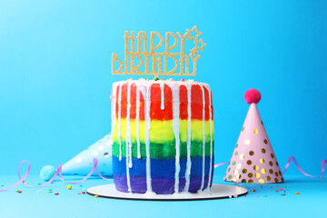 Delicious rainbow birthday cake with topper, sprinkles and party hats on light blue background
