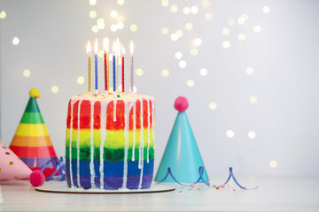 Delicious rainbow birthday cake with burning candles, streamer, sprinkles and party hats on white...
