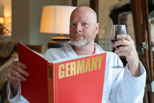 Bald man with beard drinking a glass of red wine and holding a large book on the country of Germany. The person is wearing a bathrobe. 