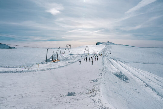 A snowy ski slope at Zermatt ski resort in Switzerland, featuring skiers on a groomed path, ski lift structures, expansive snowfields, and a cloudy sky.