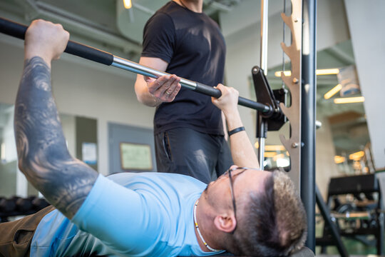 Hardworking athlete lifting heavy barbell with support from personal trainer in gym. Sports for upper body strength, discipline in physical fitness exercises, structured training and body building