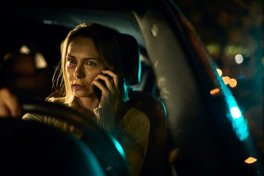 A young Caucasian woman, bearing the distressing signs of domestic abuse with prominent bruises and blackened eyes, speaks on her phone as she navigates her car through the nighttime streets.