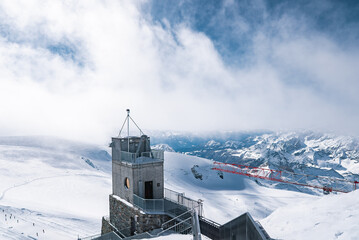 Snowy alpine landscape with a stone and metal observation tower, skiers on trails, a red crane, and mountain ranges in Glacier Paradise in Zermatt.