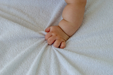 Close-up of a baby's tiny hand grasping a soft white bed sheet, showing delicate fingers and chubby...