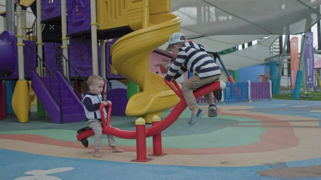Two kids are playfully enjoying their time on a colorful seesaw at the playground, under a shaded structure