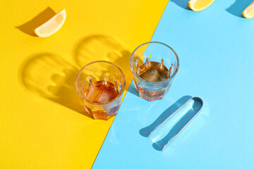 Glasses of rum with ice cubes, tongs and lemon slices on colorful background