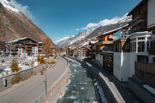View of Zermatt, Switzerland, featuring a clear stream, alpine style buildings, snow capped mountains, a clean road, and scattered trees.