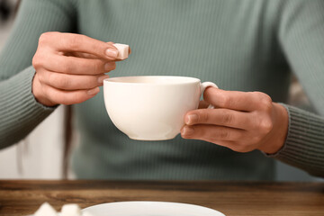 Woman adding lump of sugar into tea at table in kitchen. Closeup