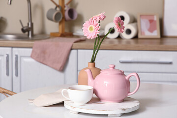 Pink teapot with cup of tea and beautiful flowers on table in kitchen
