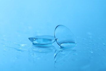 Pair of contact lenses and water drops on light blue mirror surface, closeup