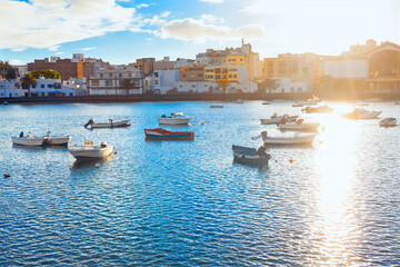 Fototapeta premium Sunlit coastal scene in Arrecife, Lanzarote, with small boats floating on calm waters. City buildings reflect the island's charm. Golden light adds warmth, creating a peaceful maritime atmosphere