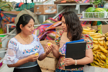 Two women, dressed in vibrant traditional attire, thoughtfully examine fresh fruits amidst a lively...
