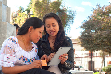 In a vibrant outdoor setting filled with sunlight, two women are actively engaging with a tablet, exchanging ideas