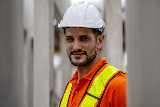 A male civil engineer in orange safety suit conducts a quality check of precast or cement floor products in a factory. A beard factory worker with hardhat stands next to cement knockdown wall