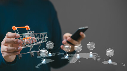 A person holding a smartphone while shopping online, featuring a miniature shopping cart and...